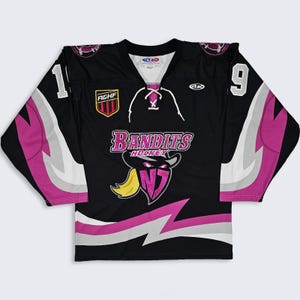 Op de afbeelding: Een zwart ijshockey-shirt met roze en zilveren bliksemschicht-accenten op de mouwen en de zoom. De voorkant toont een Bandits Hockey-logo met een gemaskerd figuur, de tekst AGHF en het nummer 19 op de mouwen. Het heeft een vetersluiting bij de kraag.