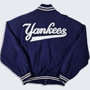 Könnte beinhalten: Eine marineblaue Baseballjacke mit weißen, gestreiften Rippen an Kragen, Ärmelbündchen und Saum. Die Rückseite der Jacke zeigt ein weiß gesticktes "Yankees"-Logo.