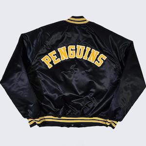 Puede incluir: Chaqueta de satén negro con la palabra "PENGUINS" arqueada en la espalda en letras amarillas y blancas. La chaqueta tiene ribetes a rayas amarillas y blancas en el cuello, los puños y el dobladillo. Una chaqueta clásica de temática deportiva.
