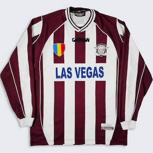 May include: A long-sleeved soccer jersey with vertical maroon and white stripes. The jersey features the text "LAS VEGAS" in blue, a crest, and the brand name "GARMAN". The jersey has a V-neck collar and white cuffs.