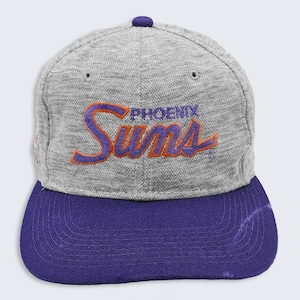 Puede incluir: Gorra de béisbol gris con visera morada y botón morado. La gorra presenta el texto bordado "PHOENIX Suns" en naranja y morado. Un accesorio deportivo.