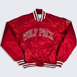 May include: A red satin varsity jacket featuring the text WOLF PACK in white lettering across the chest. The jacket has a snap-button front closure, a ribbed collar, and ribbed cuffs with white stripes. A small white logo is on the left sleeve.