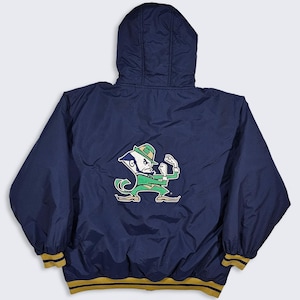 Puede incluir: Una chaqueta azul marino con capucha, mangas largas, puños elásticos y un dobladillo con rayas doradas y negras. La parte posterior muestra un gran gráfico bordado de un personaje de duende luchador con un sombrero verde.