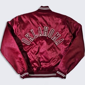 May include: A maroon satin bomber jacket with white ribbed trim and the word "OKLAHOMA" embroidered on the back in white.
