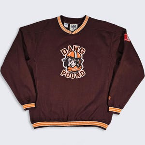 May include: A brown long-sleeve sweatshirt with orange and white striped trim at the collar, cuffs, and hem. The front features a graphic of a dog wearing a helmet and holding a football, with the text "DAWG POUND" arched around the illustration.
