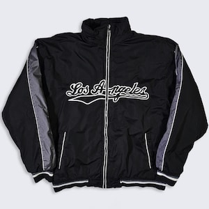 Puede incluir: Chaqueta negra con cremallera y la inscripción "Los Angeles" en blanco en el pecho. La chaqueta presenta paneles grises en las mangas y ribetes blancos en los puños y el dobladillo. Tiene un cuello alto y una cremallera completa.
