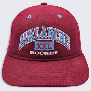 May include: Maroon baseball cap with a curved brim. The cap features the word "AVALANCHE" in blue and white embroidery, arched across the front. Below, a blue rectangle with "XXL" and "HOCKEY" in white complete the design. Two small blue eyelets are on the crown.