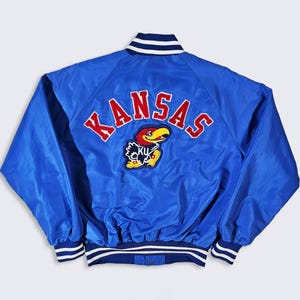 May include: A royal blue satin jacket with the word "KANSAS" in red block letters across the back. A patch of a cartoon bird in red, yellow, and black is below the word. The jacket has white and blue striped trim on the collar, cuffs, and waist.