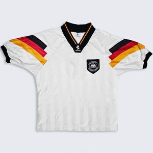 May include: White soccer jersey with black, red, and yellow accents. The jersey features a V-neck collar with black trim and the German national emblem on the chest. The sleeves have stripes of black, red, and yellow. The jersey has vertical stripes.