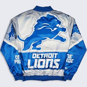 Peut inclure: Un blouson bomber en satin bleu et argenté arborant un grand logo des Detroit Lions dans le dos. Le texte "DETROIT LIONS" est imprimé en blanc sous le motif du lion, avec l'inscription "ONE PRIDE" sur les manches et un col et ourlet côtelés.