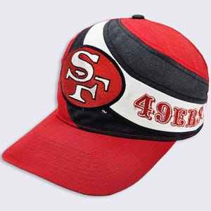 May include: A red, black, and white baseball cap with the San Francisco 49ers logo and "49ers" embroidered on the front.