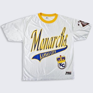 Peut inclure: T-shirt blanc avec col et bord des manches jaunes. Le t-shirt présente le mot "Monarchs" en écriture jaune avec un contour bleu, et "Kansas City" en bleu. Un logo de couronne avec "KC" est en bas à droite.