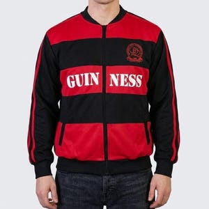 K&ouml;nnte beinhalten: Schwarz-rote Jacke mit Rei&szlig;verschluss und dem Wort "GUINNESS" in wei&szlig;en Blockbuchstaben auf der Brust. Die Jacke hat einen schwarzen Kragen, &Auml;rmel und R&uuml;cken mit roten Eins&auml;tzen auf der Vorderseite. Ein kleines Emblem ist sichtbar.