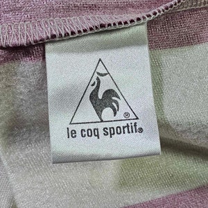 Puede incluir: Una etiqueta blanca con un logotipo de gallo negro dentro de un tri&aacute;ngulo y el texto "le coq sportif" en negro.