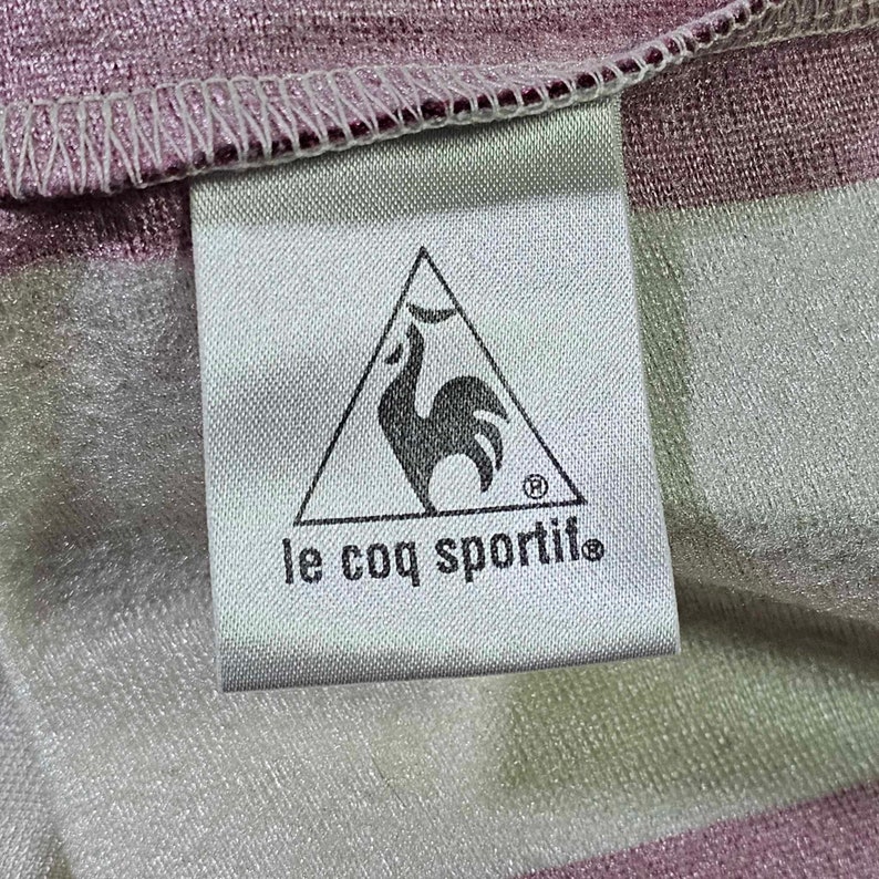 Puede incluir: Una etiqueta blanca con un logotipo de gallo negro dentro de un tri&aacute;ngulo y el texto "le coq sportif" en negro.