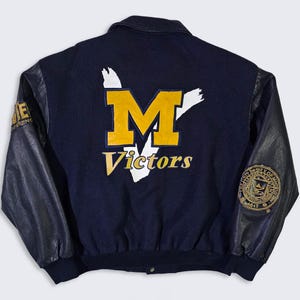 May include: Navy blue varsity jacket with black leather sleeves. The back features a large yellow "M" logo with the word "Victors" underneath. The left sleeve has a patch that says "GAME CONFERENCE". The right sleeve has a circular patch with text.