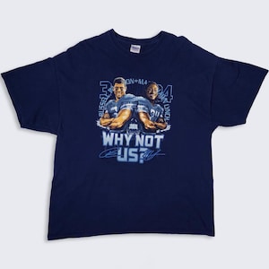 May include: Navy blue t-shirt featuring a graphic of two football players with the text "WHY NOT US?". The players are depicted with their arms crossed, wearing football jerseys. The shirt has a classic crew neck design.