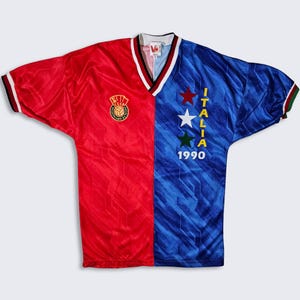 Pode incluir: Uma camisa de futebol vermelha e azul com o texto "ITALIA 1990" e um design de estrelas no lado direito. O lado esquerdo da camisa tem um patch com o texto "CCCP" e uma bola de futebol.