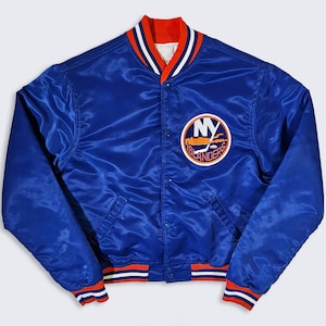 May include: A blue satin jacket with the New York Islanders logo on the chest. The jacket has a button-up front, a red and white striped collar, cuffs, and waistband. The logo features the letters "NY" and a hockey stick.