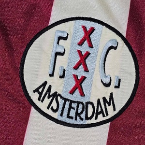 Puede incluir: Un parche bordado blanco y rojo con el texto "F.C. Amsterdam" y tres "X" rojas en una l&iacute;nea vertical.