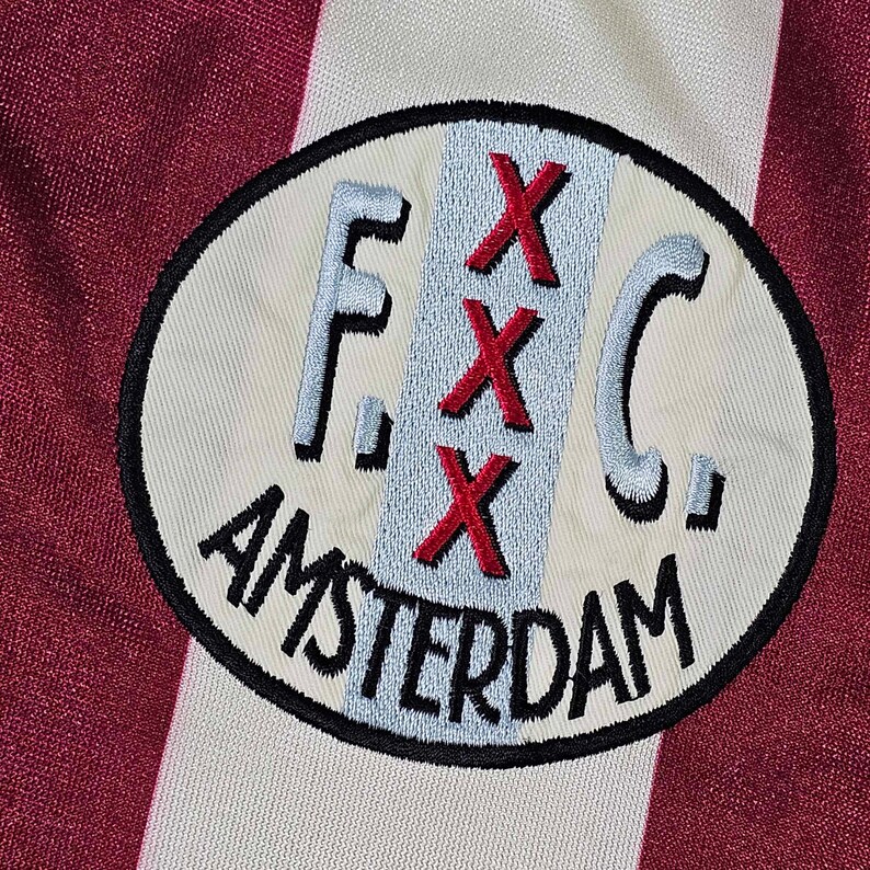 Puede incluir: Un parche bordado blanco y rojo con el texto "F.C. Amsterdam" y tres "X" rojas en una l&iacute;nea vertical.