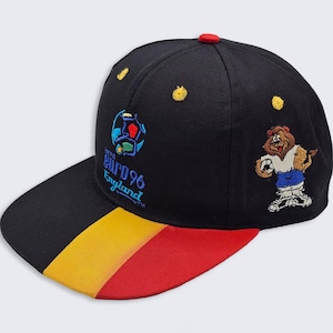 Puede incluir: Gorra de béisbol negra con visera plana, con el logo Euro 96 England y un león de dibujos animados sosteniendo un balón de fútbol. La visera tiene rayas amarillas, rojas y negras. La gorra tiene un botón rojo en la parte superior.