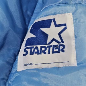 May include: Close-up of a light blue satin jacket with a white label. The label features the word "STARTER" in blue block letters, a blue star, and a space for a name.