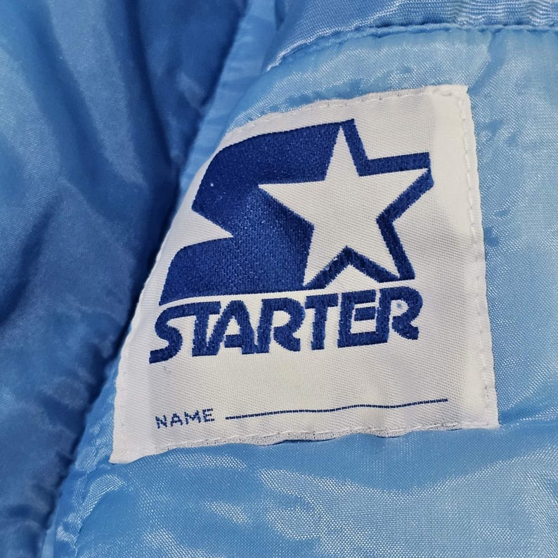 May include: Close-up of a light blue satin jacket with a white label. The label features the word "STARTER" in blue block letters, a blue star, and a space for a name.