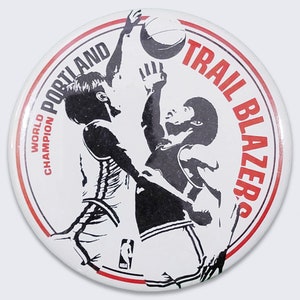 Puede incluir: Un botón blanco con un borde rojo que presenta una silueta en blanco y negro de dos jugadores de baloncesto en acción. El texto "PORTLAND TRAIL BLAZERS" está escrito en un círculo rojo alrededor de los jugadores. El texto "WORLD CHAMPION" está escrito en un rectángulo rojo a la izquierda de los jugadores.