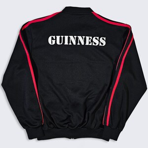 K&ouml;nnte beinhalten: Schwarze Trainingsjacke mit roten Streifen an &Auml;rmeln und Schultern. Der Schriftzug "GUINNESS" ist in Wei&szlig; auf dem R&uuml;cken aufgedruckt. Die Jacke hat einen gerippten Kragen und B&uuml;ndchen. Klassische Sportbekleidung.