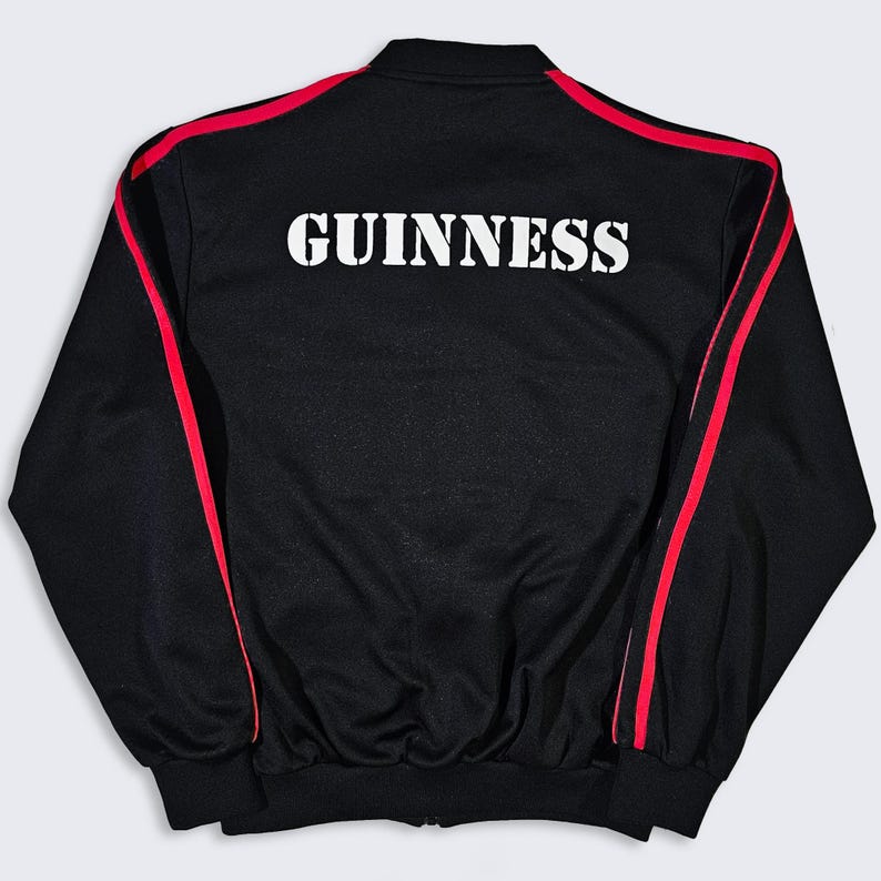 K&ouml;nnte beinhalten: Schwarze Trainingsjacke mit roten Streifen an &Auml;rmeln und Schultern. Der Schriftzug "GUINNESS" ist in Wei&szlig; auf dem R&uuml;cken aufgedruckt. Die Jacke hat einen gerippten Kragen und B&uuml;ndchen. Klassische Sportbekleidung.