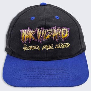 Puede incluir: Gorra de béisbol negra y azul con el texto "WAR WIZARD" bordado en oro y morado. La gorra tiene una visera curva y un botón en la parte superior. El texto "UNDER ROCK RACING" está bordado debajo.