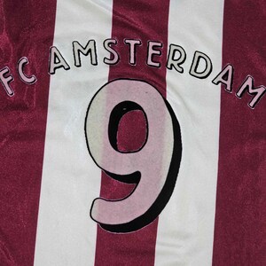 Puede incluir: Una camiseta de f&uacute;tbol a rayas blancas y burdeos con el texto "FC AMSTERDAM" y el n&uacute;mero "9" en la parte delantera.