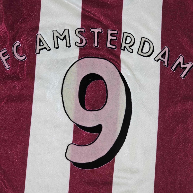Puede incluir: Una camiseta de f&uacute;tbol a rayas blancas y burdeos con el texto "FC AMSTERDAM" y el n&uacute;mero "9" en la parte delantera.