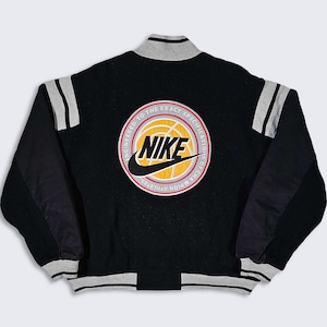 May include: Black varsity jacket with a large embroidered Nike logo on the back. The logo features a basketball and the text "Engineered to the Exact Specification of Champion Athletes".