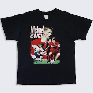 May include: Black t-shirt with a graphic print of Michael Owen, a famous soccer player, in a red and white soccer uniform. The text "Michael Owen" is printed in white on the shirt.
