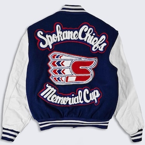 Puede incluir: Una chaqueta de béisbol azul y blanca con el texto "Spokane Chiefs Memorial Cup" en la espalda. La chaqueta tiene mangas blancas y un cuerpo azul con un logotipo rojo y blanco.