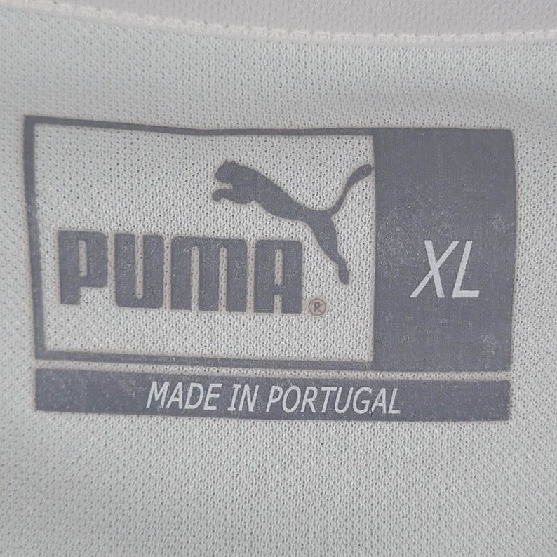 K&ouml;nnte beinhalten: Ein graues Puma-Logo mit dem Wort "Puma" und einem eingetragenen Warenzeichen. Die Gr&ouml;&szlig;e "XL" ist unter dem Logo gedruckt. Der Text "Made in Portugal" ist unter der Gr&ouml;&szlig;e gedruckt.