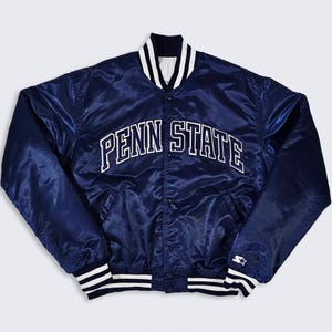 Puede incluir: Chaqueta de satén azul marino con el texto "PENN STATE" en letras blancas en el pecho. La chaqueta tiene una botonadura frontal, puños y cuello a rayas blancas y negras, y un logotipo de Starter en la manga.