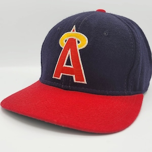 Puede incluir: Una gorra de béisbol azul marino con visera roja. La parte delantera de la gorra presenta una 'A' bordada en blanco con un halo amarillo encima.