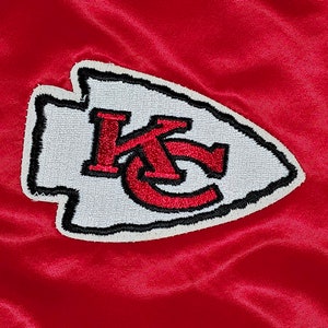 May include: A white and red embroidered patch with the letters "KC" in a stylized arrowhead shape. The patch is on a red background.