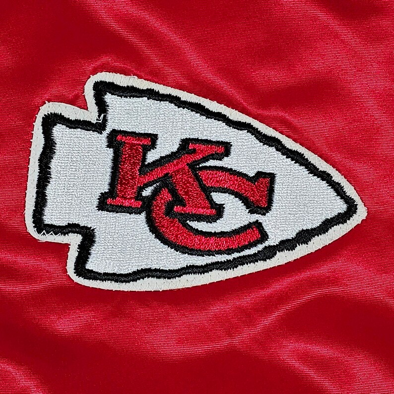 May include: A white and red embroidered patch with the letters "KC" in a stylized arrowhead shape. The patch is on a red background.