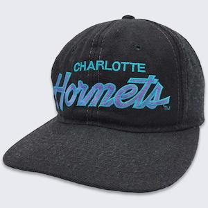 以下が含まれることがあります： 黒のベースボールキャップに、ターコイズとパープルの刺繍で「CHARLOTTE Hornets」のロゴが入っています