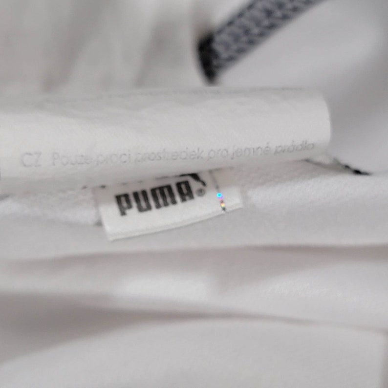 K&ouml;nnte beinhalten: Nahaufnahme eines wei&szlig;en Puma-Logos mit dem Text "CZ Pouze proci prostředek pro jemn&eacute; pr&aacute;dlo" auf Tschechisch.