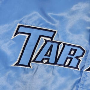 May include: Close-up of a light blue satin jacket with the embroidered word "TAR" in navy blue and white. The jacket has a shiny, smooth texture and a classic athletic style.