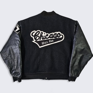 May include: Black varsity jacket with leather sleeves. The back features a white embroidered "Chicago White Sox" logo. A patch is on the left sleeve. The jacket has a black ribbed collar and cuffs.