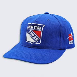 May include: A royal blue baseball cap featuring the New York Rangers logo on the front. The logo is a shield shape with the words "NEW YORK RANGERS" and a red and white design. The cap also has the "SPORTS SPECIALISTS" logo on the side.