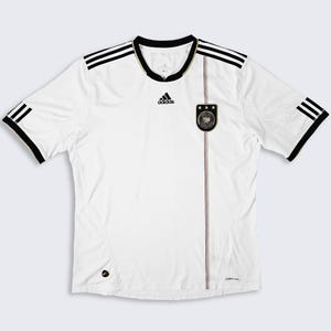 May include: White soccer jersey with black and gold accents. The jersey features the Adidas logo, black stripes on the shoulders and sleeves, and a German national team crest. The jersey has a crew neck and short sleeves.