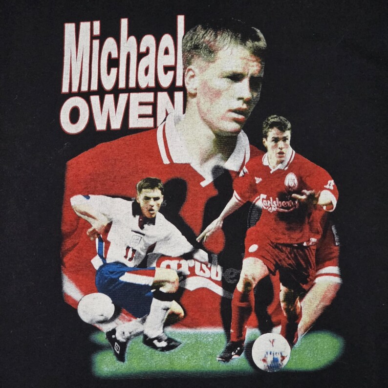 May include: A black t-shirt with a red, white, and blue graphic of Michael Owen, a famous soccer player. The graphic features Owen in a red soccer jersey with the text "Carlsberg" on the front. The text "Michael Owen" is printed in white on the shirt.