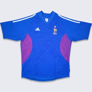 Peut inclure: Un maillot de football bleu avec des accents blancs et violets. Le maillot porte le logo Adidas et l'écusson de l'équipe nationale française avec un coq et les lettres FFF.